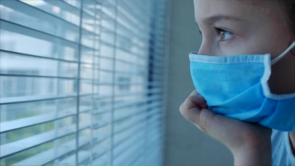 Ein kleines Kind oder kleines Mädchen in einer medizinischen Schutzmaske schaut hinter einem Vorhang aus dem Fenster, mit einem traurig enttäuschten Gesicht. Mädchen mit medizinischer Maske. Pandemie, covid - 19 — Stockvideo