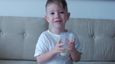 Evdeki kanepede oturup süt içen tatlı bebek. Yavaş çekim su içen küçük çocuk. Yakın plan. Komik küçük çocuk bir bardak su içiyor..