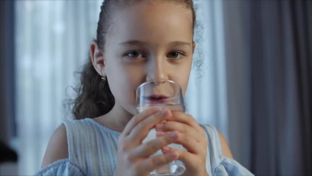 Carina bambina che beve un bicchiere d'acqua seduta sul divano di casa. Ragazzino al rallentatore che beve acqua. Primo piano. Ritratto Funny little Child beve una tazza d'acqua. — Video Stock