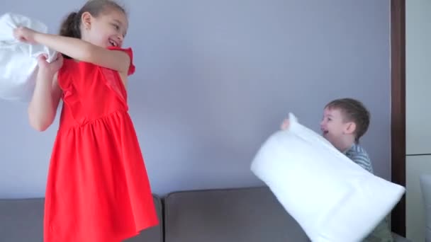 Щаслива молодша сестра з маленьким молодшим братом насолоджуються смішною бійкою на ліжку, брат і сестра організували бійку подушками на ліжку в спальні.. — стокове відео