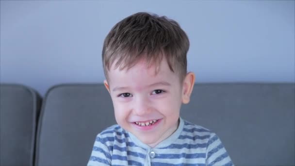 Portrét Vtipný chlapeček, usmívající se na kameru, sedí doma na gauči, roztomilé dítě ve školce s hezkým obličejem sedícím na pohovce. Koncept Happy Childhood. — Stock video