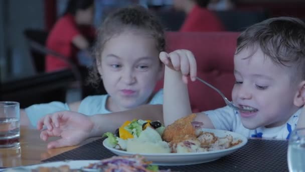 Portret van een kind jongen en meisje eet aardappelpuree met salade, tomaten, broccoli en rode saus in een lichte huiskamer. Een klein kind draait spaghetti op een vork en eet maaltijd.. — Stockvideo