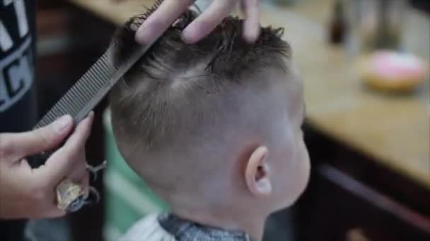 Coupe de cheveux d'enfants dans le salon au moment de l'épidémie de coronavirus. Le petit garçon se fait couper les cheveux. Coiffure d'enfant. Styliste dans un masque de protection de covid-19 fait une coiffure élégante. — Video