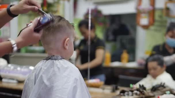 O rapazinho corta o cabelo. Estilo de cabelo das crianças. Estilista em uma máscara protetora de covid-19 faz um penteado elegante. Corte de cabelo das crianças no salão no momento da epidemia de coronavírus. — Vídeo de Stock