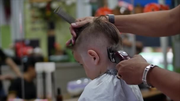 Le petit garçon se fait couper les cheveux. Coiffure d'enfant. Styliste dans un masque de protection de covid-19 fait une coiffure élégante. Coupe de cheveux d'enfants dans le salon au moment de l'épidémie de coronavirus. — Video