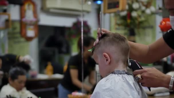 Kleiner Junge bekommt einen Haarschnitt. Kinderhaarstyling. Stylist in einer Schutzmaske von covid-19 macht eine stylische Frisur. Kinderfrisur im Salon zur Zeit der Coronavirus-Epidemie. — Stockvideo