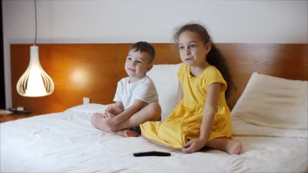 Portret uroczych dzieci oglądających wieczorem telewizję w domu, siedzących samotnie na dużym łóżku. Chłopiec i dziewczyna oglądanie kreskówek śmieszne i smutne, miły i przerażający w kinie domowym w żyjących — Wideo stockowe