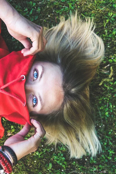 Portrait of a blonde girl with a short haircut. The background is green. Orange sweater and blue jeans.The girl with blue eyes lays on the grass.
