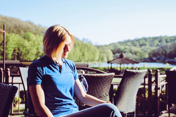 Portrait of a blonde girl with a short haircut. Background  sports ground. Dark blue t-shirt polo.The girl is sitting in a cafe.