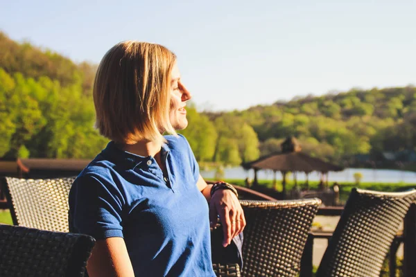 Portrait of a blonde girl with a short haircut. Background  sports ground. Dark blue t-shirt polo.The girl is sitting in a cafe.