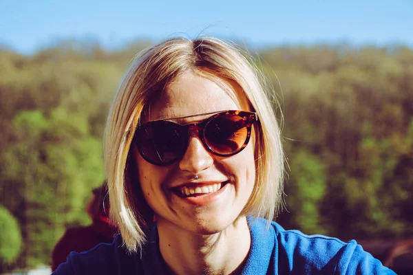 Portrait of a blonde girl with a short haircut. Background  sports ground. Dark blue t-shirt polo and brown sunglasses.