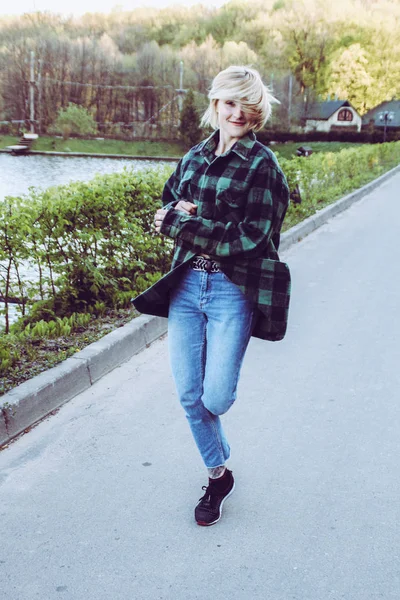 Portrait of a blonde girl with a short haircut. The background is nature.Black with a green shirt and blue jeans. Crazy girl jumped and dance.
