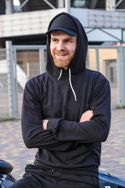 Charismatic man with red beard in a black cap and black hoodie.