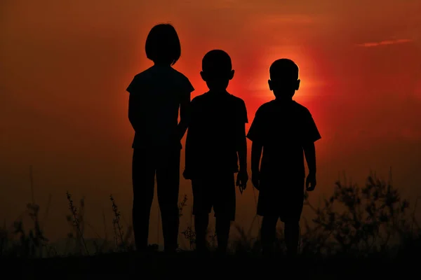 Black silhouette of three children standing together. There is a sky at sunset.