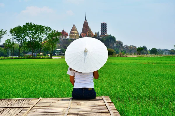 那女人坐在一座木桥上 面对着一把白伞的背 山上有草原和佛教寺庙作为背景 — 图库照片