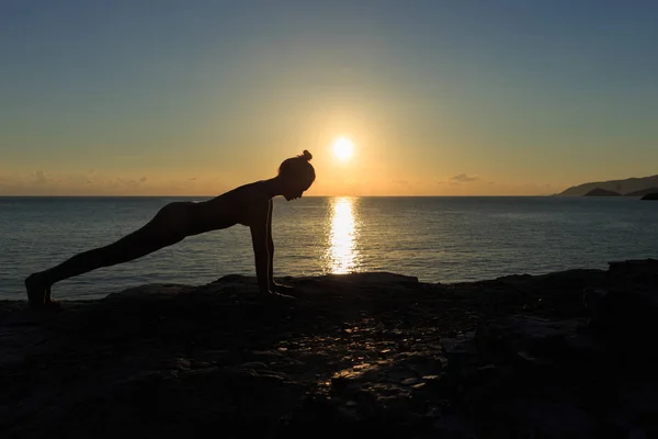 Young female silhouette doing plank exercise on a beach at sunrise