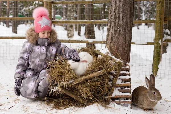Ένα χαριτωμένο κοριτσάκι σε ένα ροζ καπάκι που παίζει με ένα λευκό κουνέλι — Φωτογραφία Αρχείου