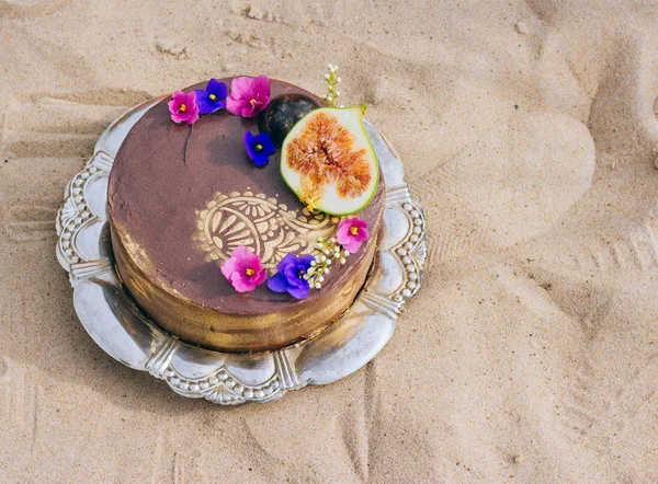 tray with chocolate cake with golden oriental glowed ornaments and pink with purple violets and figs on sandy beach