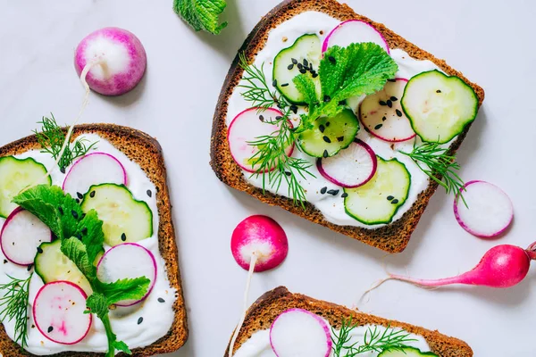 Toast with radish, cucumber and black sesame of dark bread on a light marble background