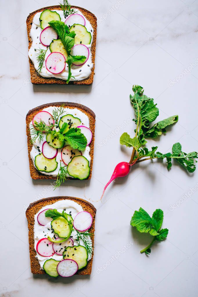 Toast with radish, cucumber and black sesame of dark bread on a light marble background