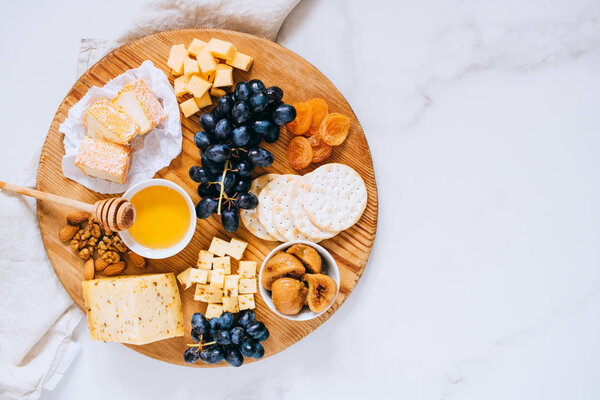 Flat lay with various types of cheese, grapes, nuts, honey and cracker in wooden board on marble