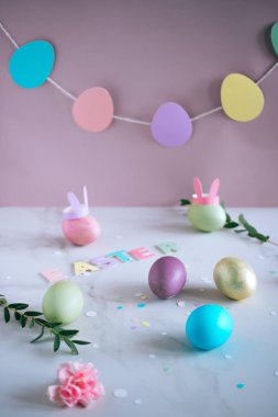 Çok renkli Paskalya yortusu yumurta, pembe, yeşil, mavi, altın tavşan kulaklı mermer arka plan, minimalizm, çiçekler. Ester garland.