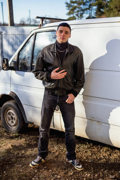 Молодой парень с криминальной внешностью в черной кожаной куртке стоит рядом со старым белым фургоном
