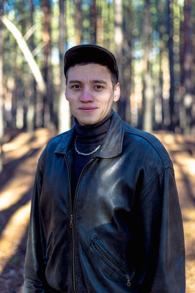 A guy in a leather jacket and cap on a road in a pine forest.