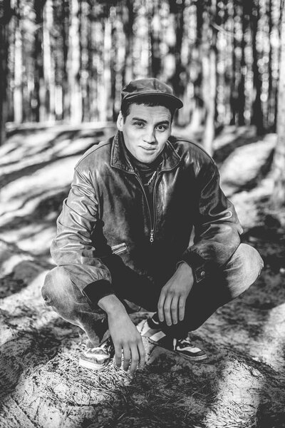 A guy in a leather jacket and cap sits on a road in a pine forest