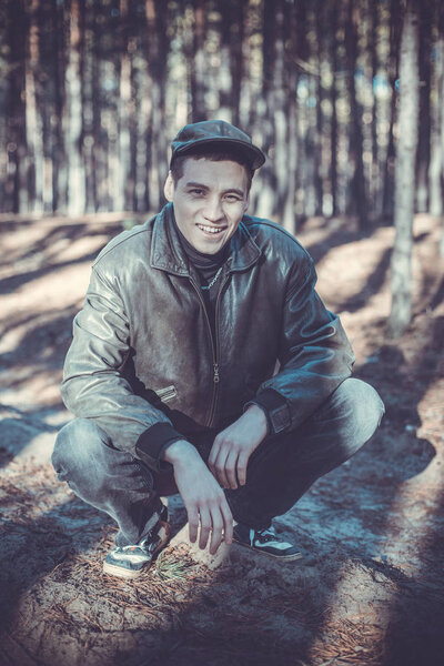 A guy in a leather jacket and cap sits on a road in a pine forest