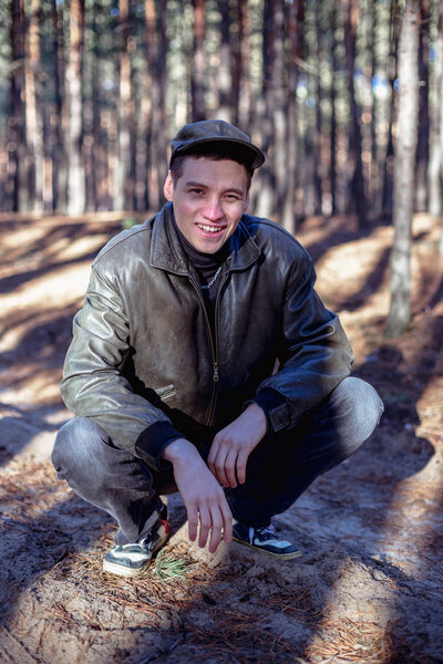 A guy in a leather jacket and cap sits on a road in a pine forest