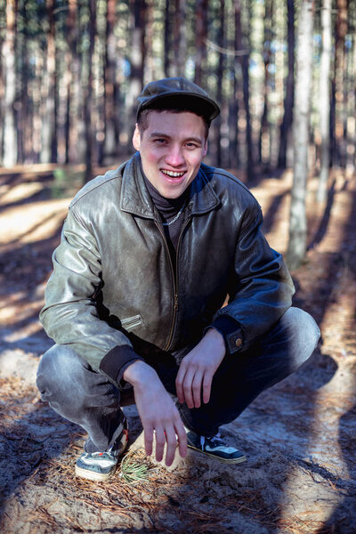 A guy in a leather jacket and cap sits on a road in a pine forest