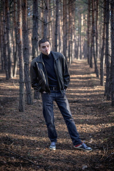 A young man of criminal appearance in a black leather jacket posing in an autumn forest