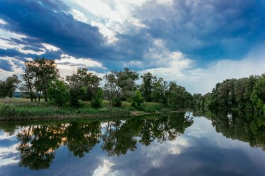 Sakin güzel doğal manzara mavi nehir, yeşil ağaçlar ve su bulutlu gökyüzü ile yansıtan. Büyülü günbatımı kırsal arazide Nehri. Doğal, vahşi manzara.