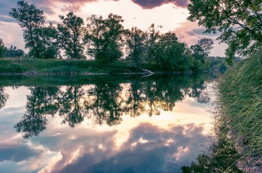 Sakin güzel doğal manzara mavi nehir, yeşil ağaçlar ve su bulutlu gökyüzü ile yansıtan. Büyülü günbatımı kırsal arazide Nehri. Doğal, vahşi manzara.