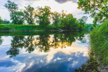 Sakin güzel doğal manzara mavi nehir, yeşil ağaçlar ve su bulutlu gökyüzü ile yansıtan. Büyülü günbatımı kırsal arazide Nehri. Doğal, vahşi manzara.