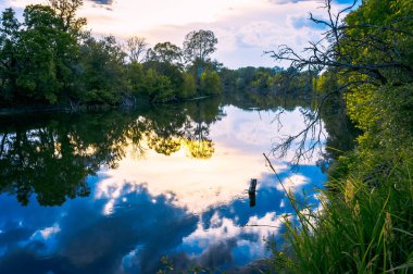 Sakin güzel doğal manzara mavi nehir, yeşil ağaçlar ve su bulutlu gökyüzü ile yansıtan. Büyülü günbatımı kırsal arazide Nehri. Doğal, vahşi manzara.