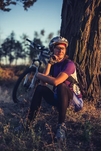 Woman in helmet sitting at sunset near her bicycle.