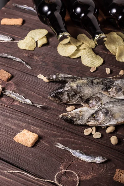 Dried fish, perch, bleak, chips, crackers, nuts and three brown glass bottles of beer on a wooden background. Studio photo.