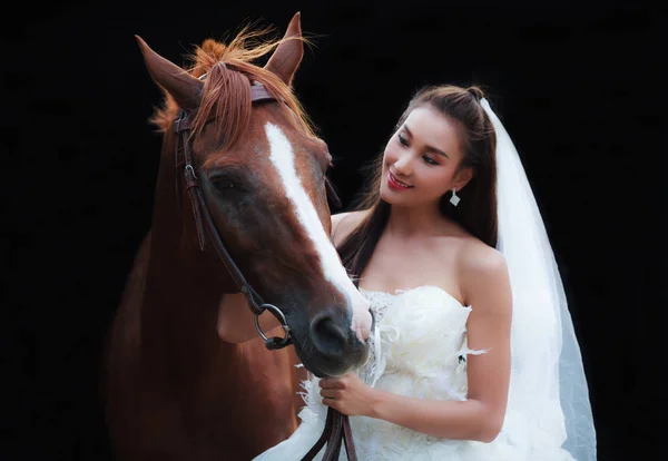 Young beautiful beauty bride in fashion white wedding costume stand with handsome horse on black background