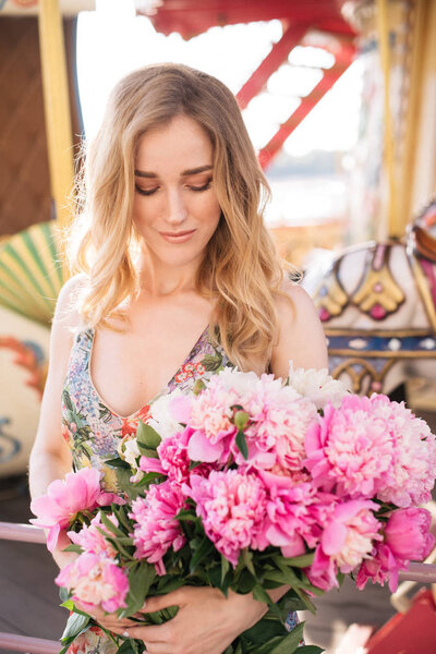 Beautiful girl in a dress with a bouquet of pink peonies in the hands near a vintage carousel