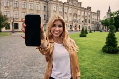 Heyecanlı genç kadın gündelik giysiler içinde modern akıllı telefon görüntüsü sergiliyor. Şehrin caddesinde yürüyüp kameraya bakıyor.