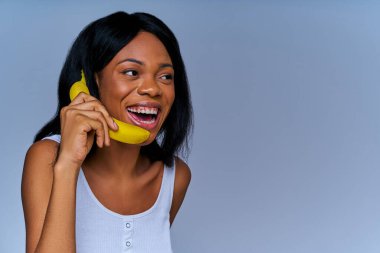 Beyaz tişörtlü, taze meyveyle oynayan gülümseyen kadın kulağına takıyor.