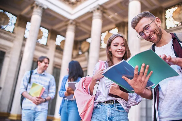 In the blurry background people are talking when in the foreground girl and guy are Viewing a notebook.