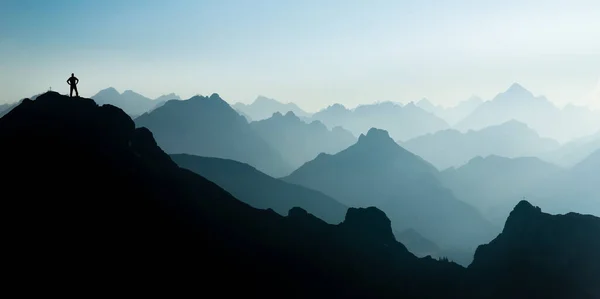 Hombre llegando a la cima después de escalar y caminar disfrutando de la libertad y mirando hacia las siluetas de las montañas panorama durante el amanecer . — Foto de Stock