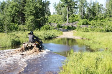 Motokros yapan bir adam ATV avlusunu su sıçratan nehir suyuna doğru sürüyor. Foy, Sudbury, Ontario, Kanada.