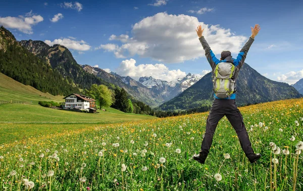 Feliz excursionista exitoso con mochila saltando con los brazos levantados en Hawkbit Flower prado. Montañas cubiertas de nieve y casa tradicional. Baviera, Alpes, Oberstdorf, Allgau, Alemania . — Foto de Stock