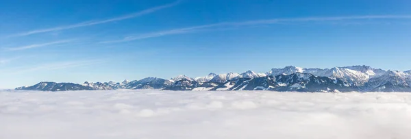 Karlı dağ sırasının fantastik panoramik görüntüsü ters sis tabakasından sarkıyor. Bulutların üzerinde güneşli mavi gökyüzü olan harika bir manzara. Ofterschwanger Borusu, Alpler, Allgau, Bavyera, Almanya. — Stok fotoğraf