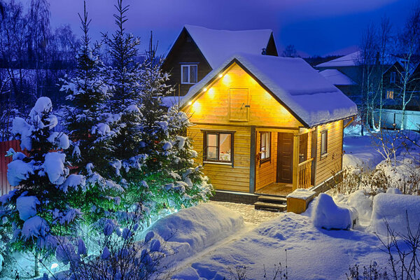 MOSCOW REGION, RUSSIA - Illuminated with spot lights Russian wooden guest house-banya (sauna) framed with spruce and birch trees covered snow in winter twilight.