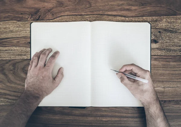 Ansicht von oben: Mann hält Stift zum Schreiben in Tagebuch oder Notizblock auf Holztisch — Stockfoto